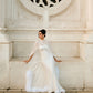 Woman in a white wedding dress and sheer silk chiffon bridal cape sitting against an ornate light stone wall, with the cape spread around her and ostrich feather trim visible along the floor-length edge.