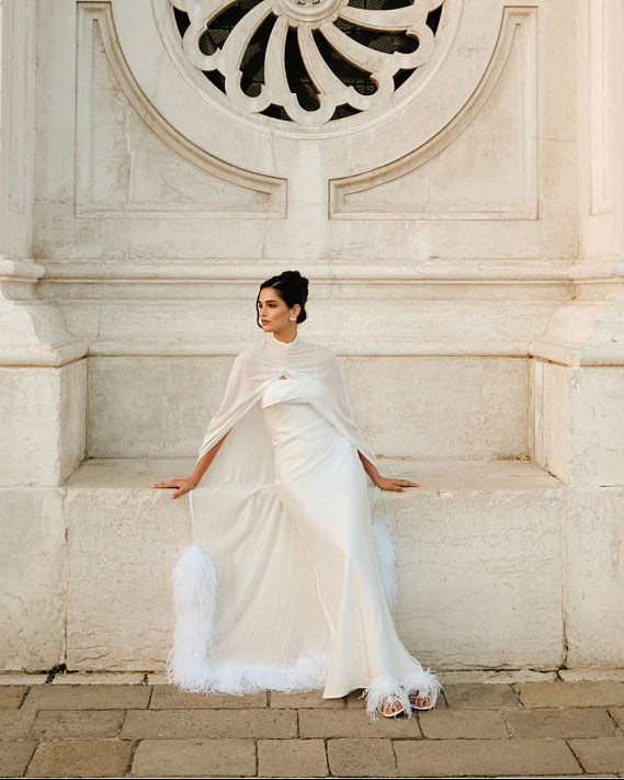 Woman in a white wedding dress and sheer silk chiffon bridal cape sitting against an ornate light stone wall, with the cape spread around her and ostrich feather trim visible along the floor-length edge.