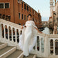 Woman in a white wedding dress wearing a floor-length silk chiffon bridal cape with ostrich feather trim, standing on a stone bridge overlooking a Venice canal and historic buildings.