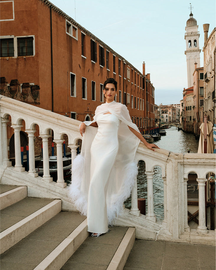 Woman in a white wedding dress wearing a floor-length silk chiffon bridal cape with ostrich feather trim, standing on a stone bridge overlooking a Venice canal and historic buildings.