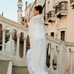 Back view of a woman wearing a floor-length sheer silk chiffon bridal cape with ostrich feather trim, standing on a stone bridge overlooking a canal in Venice.