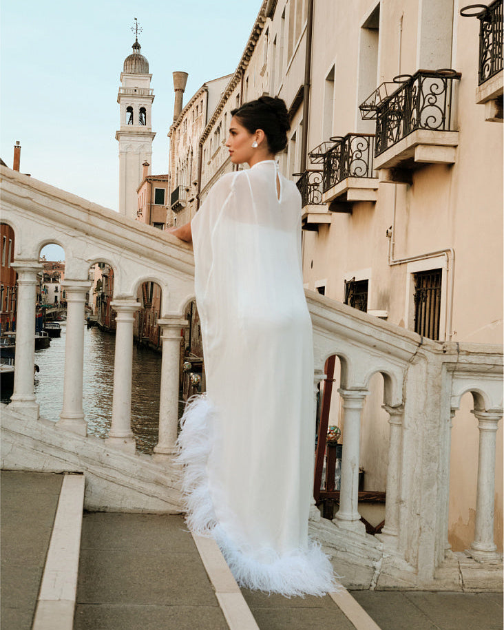 Back view of a woman wearing a floor-length sheer silk chiffon bridal cape with ostrich feather trim, standing on a stone bridge overlooking a canal in Venice.