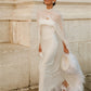 Woman walking outdoors in a white wedding dress and sheer silk chiffon bridal cape, holding a white hat; the draped cape flows behind her with ostrich feather trim along the edge.