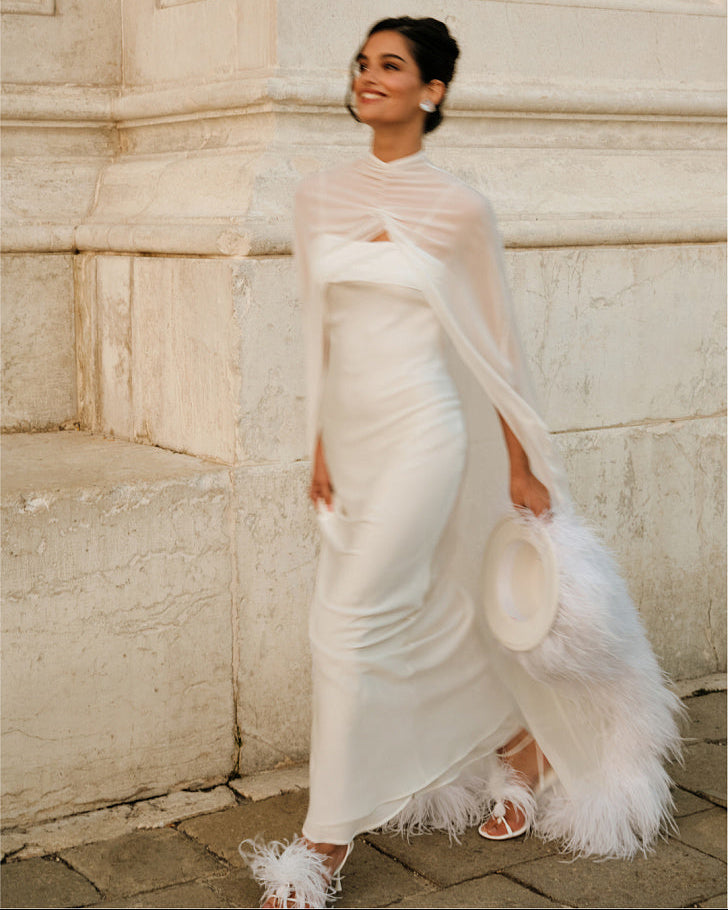 Woman walking outdoors in a white wedding dress and sheer silk chiffon bridal cape, holding a white hat; the draped cape flows behind her with ostrich feather trim along the edge.