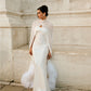 Woman wearing a floor-length white silk chiffon bridal cape over a fitted wedding dress, standing outdoors against a light stone wall; the cape is draped across the front and trimmed with ostrich feathers.