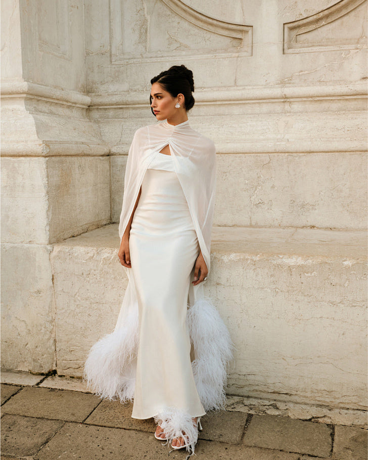 Woman wearing a floor-length white silk chiffon bridal cape over a fitted wedding dress, standing outdoors against a light stone wall; the cape is draped across the front and trimmed with ostrich feathers.