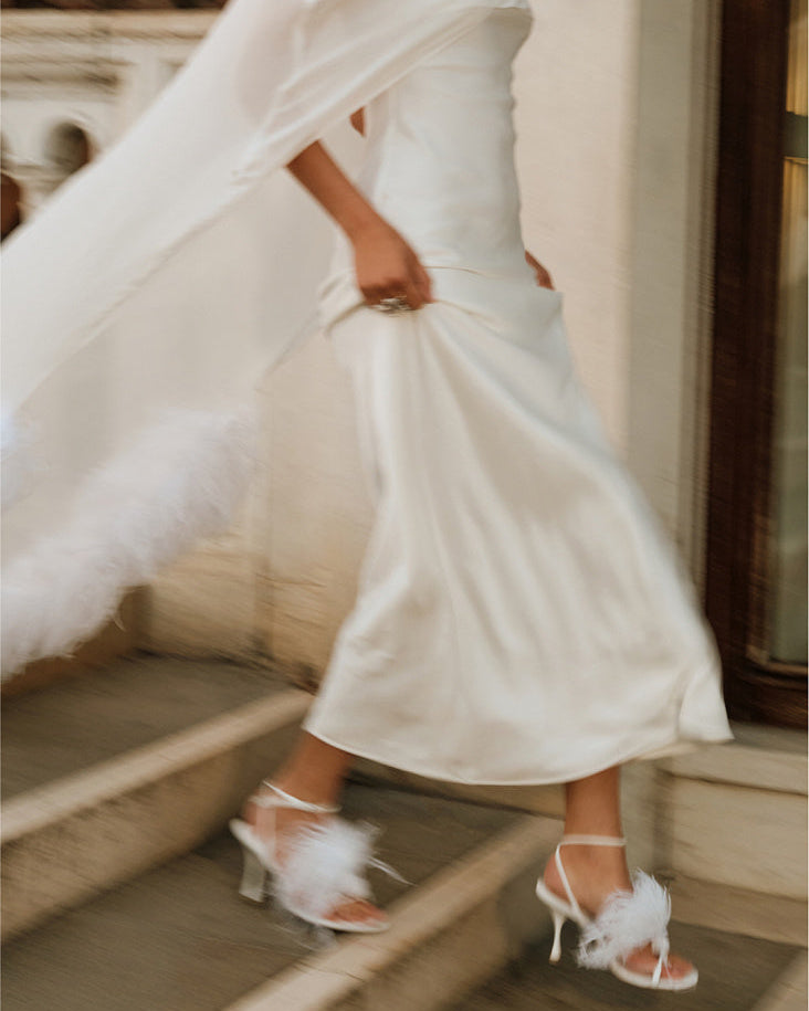 Close view of a woman walking down stone steps in a white wedding dress, with a sheer silk chiffon bridal cape flowing behind her and white feather-trimmed heels.