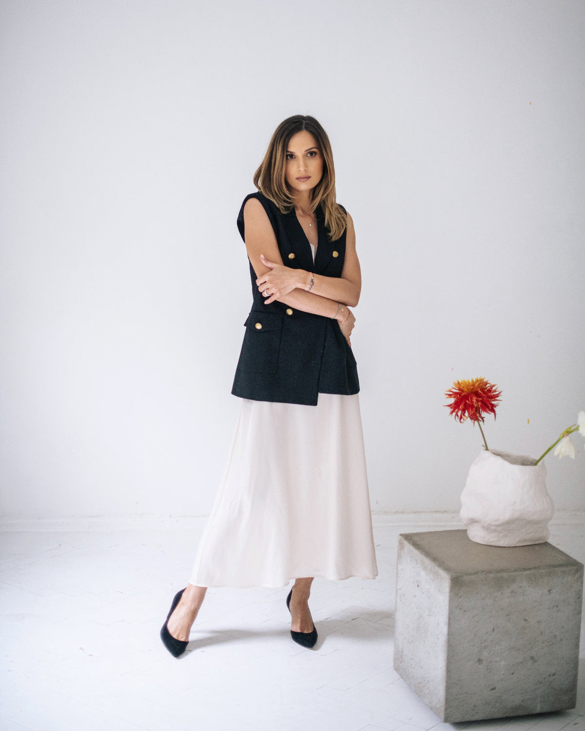 Woman in an ivory silk midi dress layered with a long black vest and worn with black heels