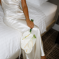 Bride wearing an ivory backless silk maxi dress sitting on a bed holding white calla lilies.
