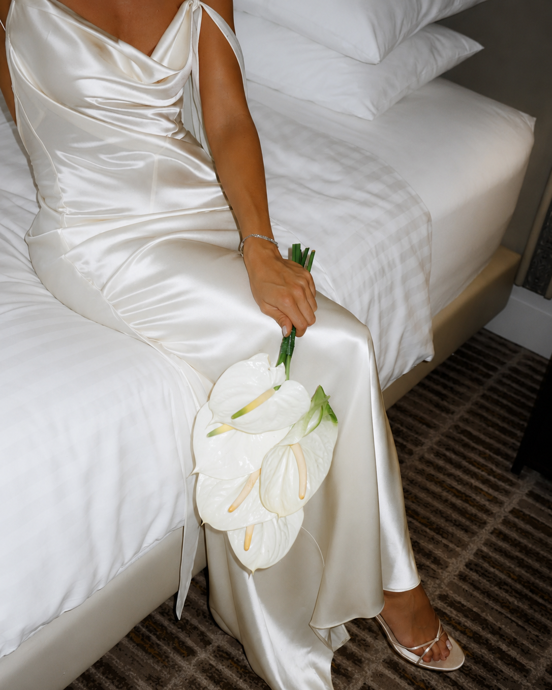 Bride wearing an ivory backless silk maxi dress sitting on a bed holding white calla lilies.
