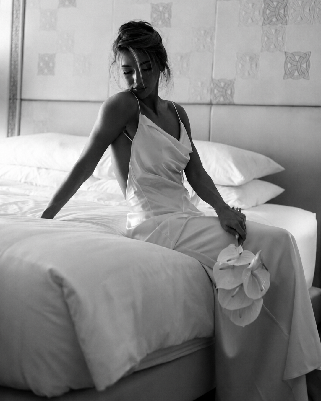 Bride wearing an ivory backless silk maxi dress sitting on a bed holding white calla lilies.