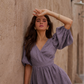 A woman wearing a lavender wrap-style dress with puff sleeves and a gathered waist, posing against a textured beige stone wall.