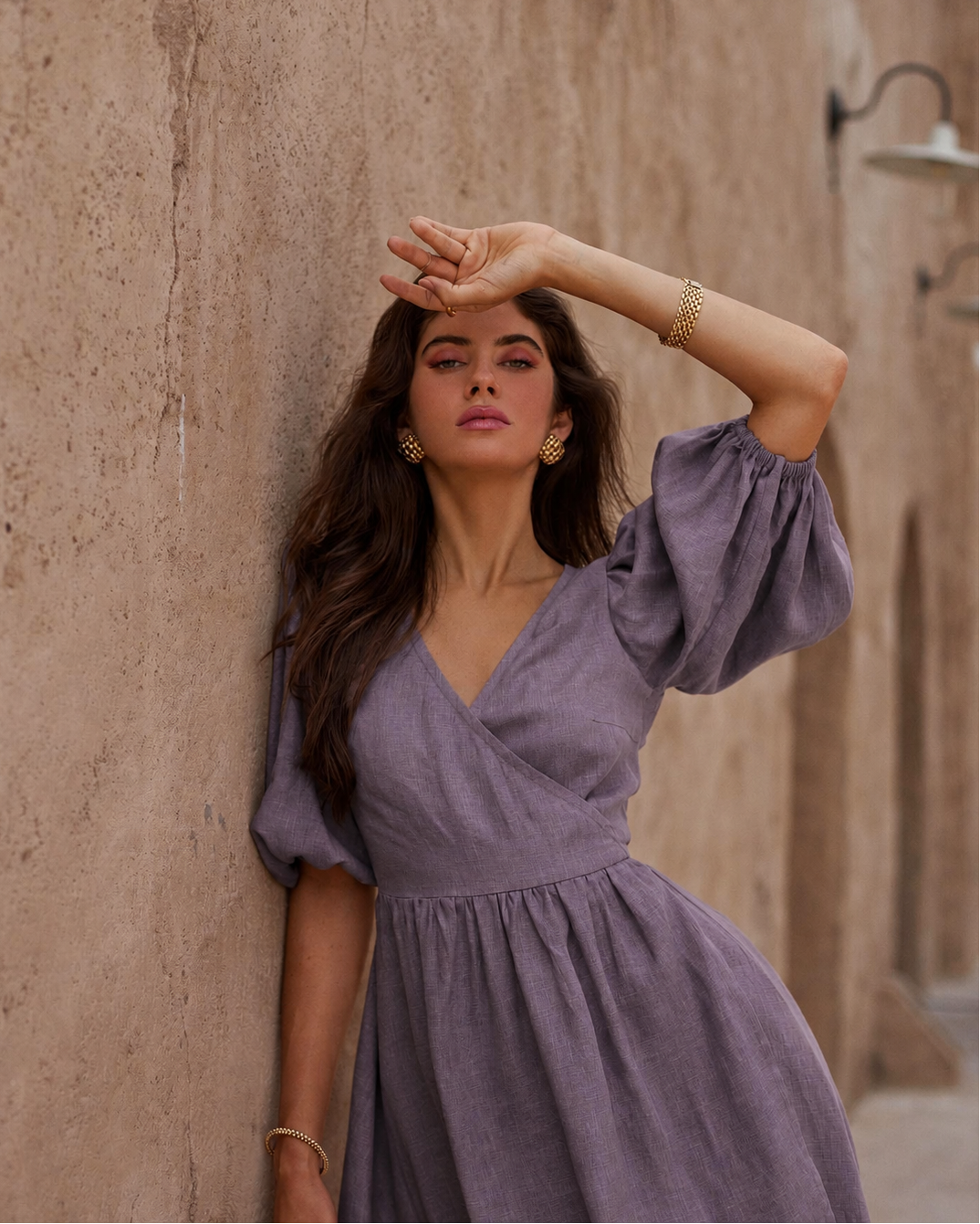 A woman wearing a lavender wrap-style dress with puff sleeves and a gathered waist, posing against a textured beige stone wall.