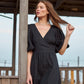 A woman wearing a black wrap-style midi dress with puff sleeves and a V-neckline, standing outdoors on a wooden boardwalk.