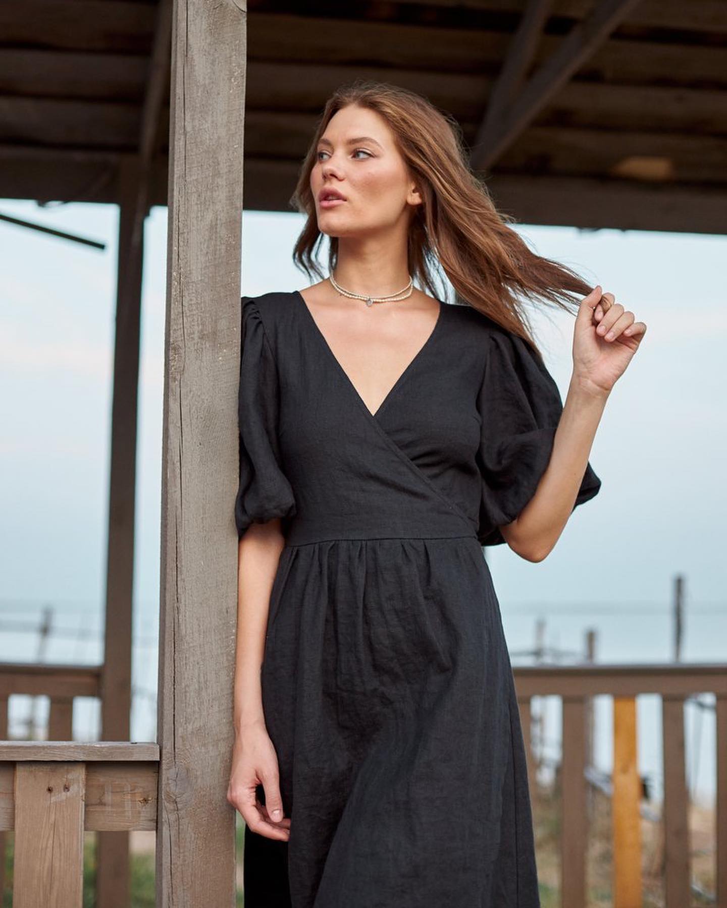 A woman wearing a black wrap-style midi dress with puff sleeves and a V-neckline, standing outdoors on a wooden boardwalk.