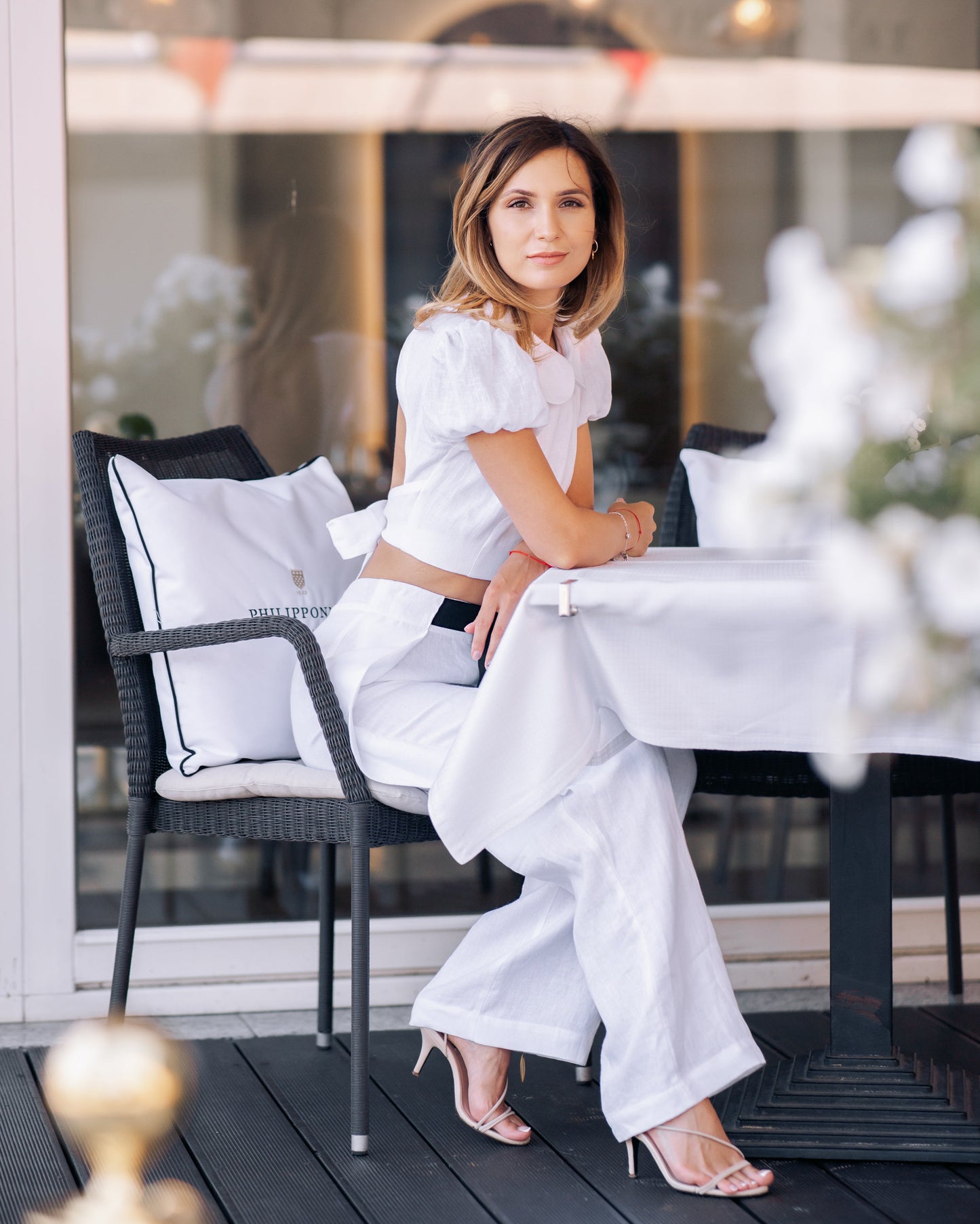 Woman wearing a white linen crop top with puff sleeves and matching wide-leg trousers, seated at an elegant outdoor city cafe.