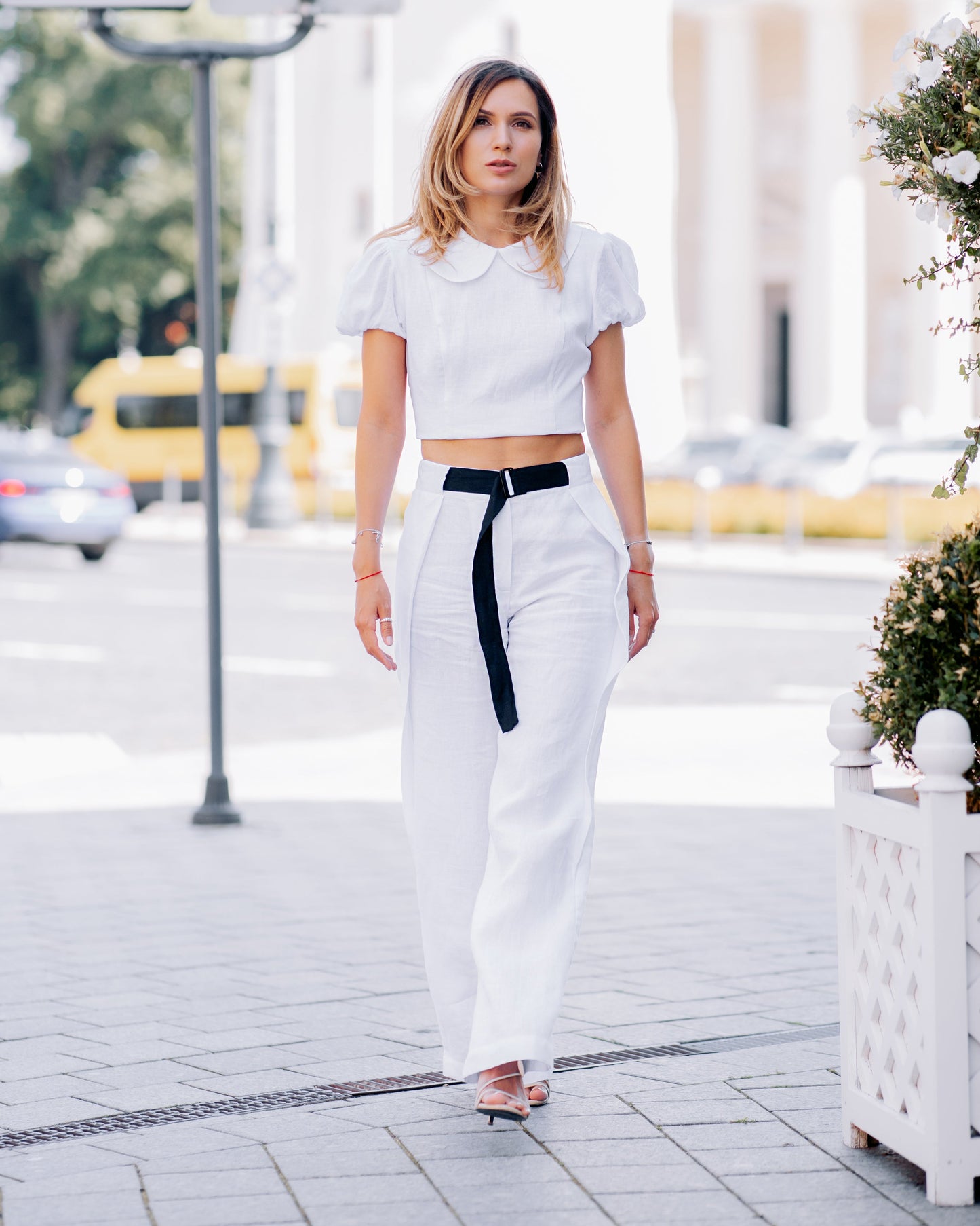 Woman walking through the city in a white linen crop top with puff sleeves, styled with white wide-leg trousers and a black belt.
