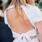 Close-up back view of a woman wearing a white linen top with a dramatic open back, puff sleeves and a bow tie at the waist.