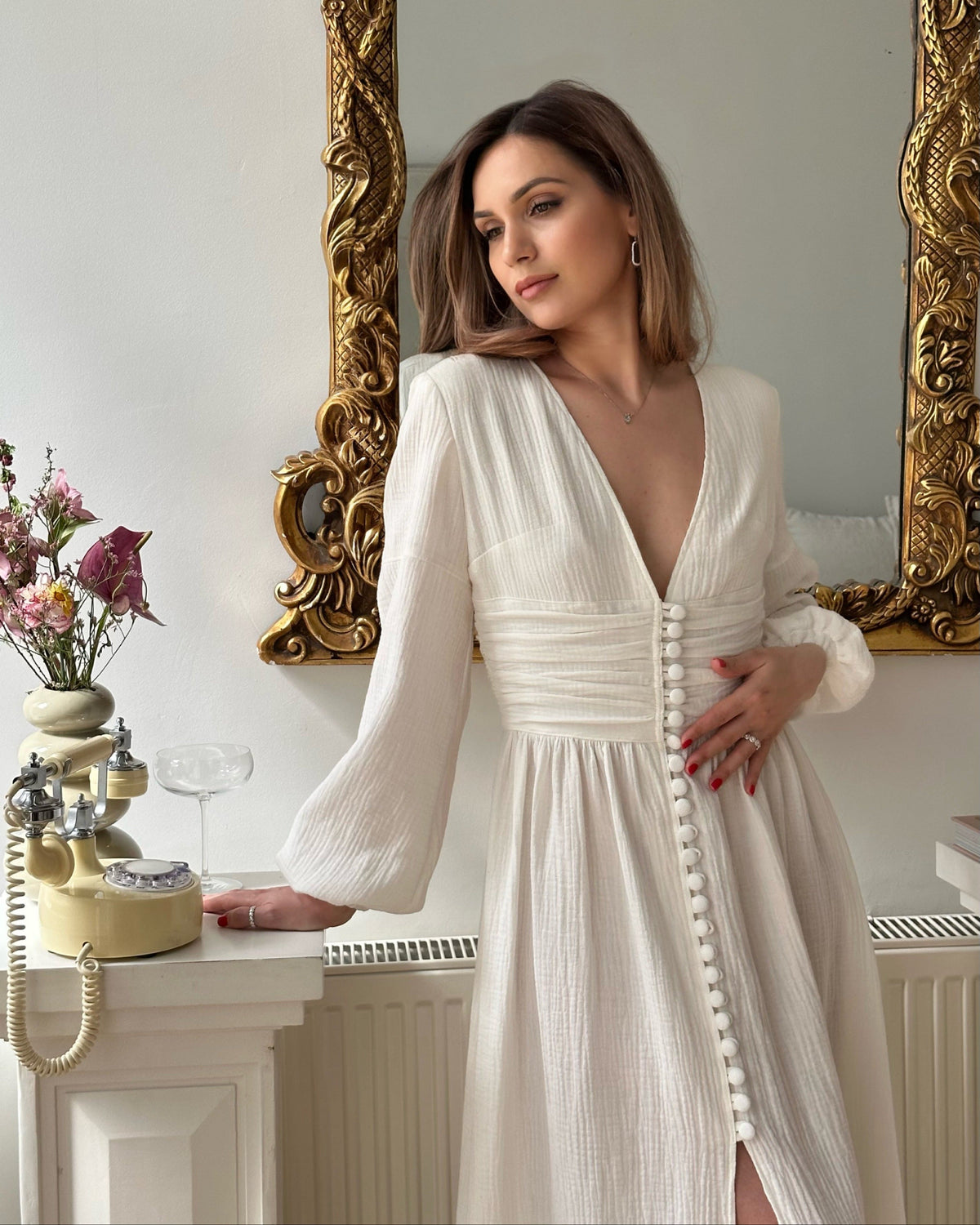 Woman wearing a white long sleeve boho wedding dress with a deep V-neck, draped waist and fabric-covered front buttons, standing beside an ornate gold mirror.
