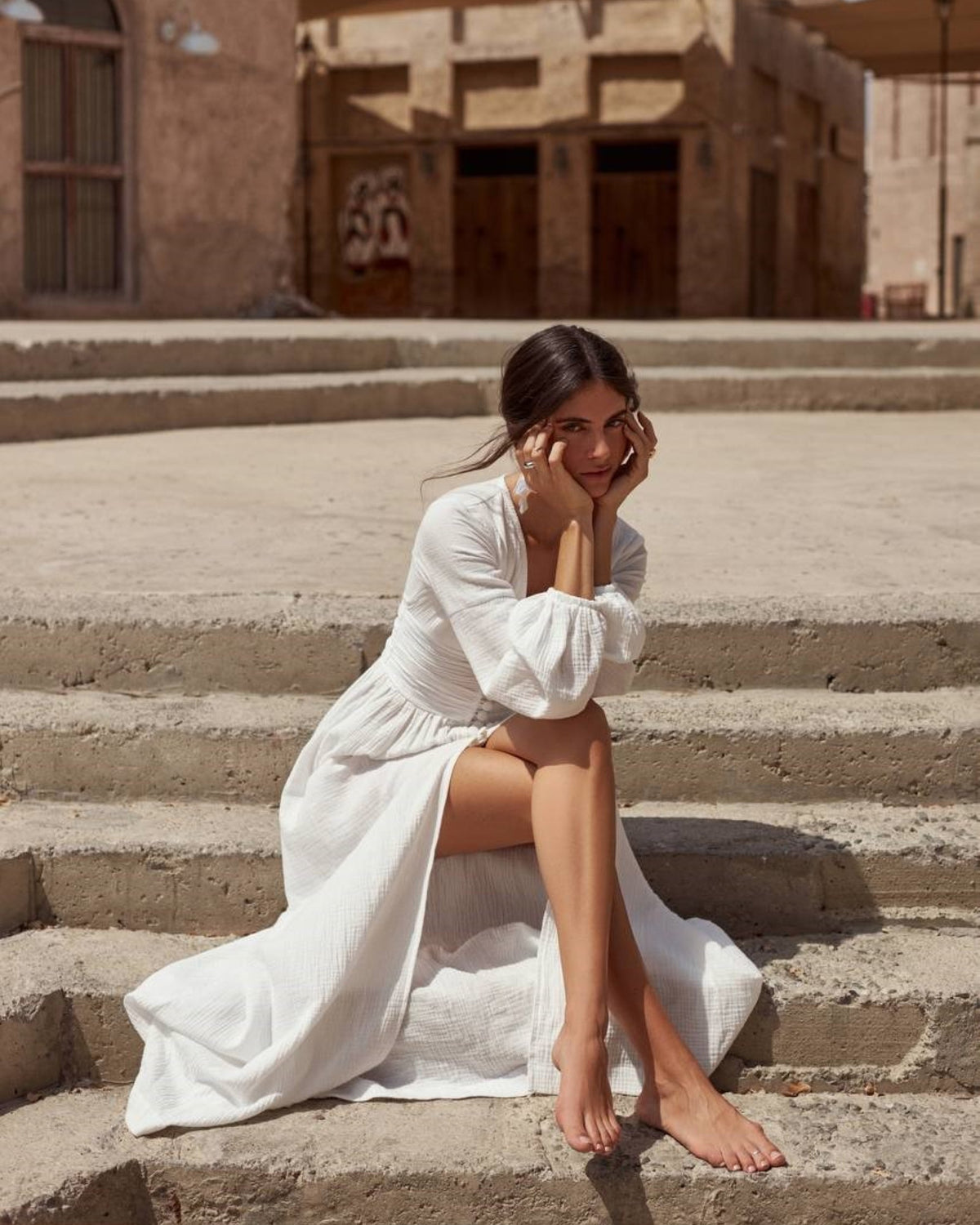 Woman wearing a white long sleeve cotton muslin wedding dress with an open button-front skirt, sitting on stone steps in the sun.