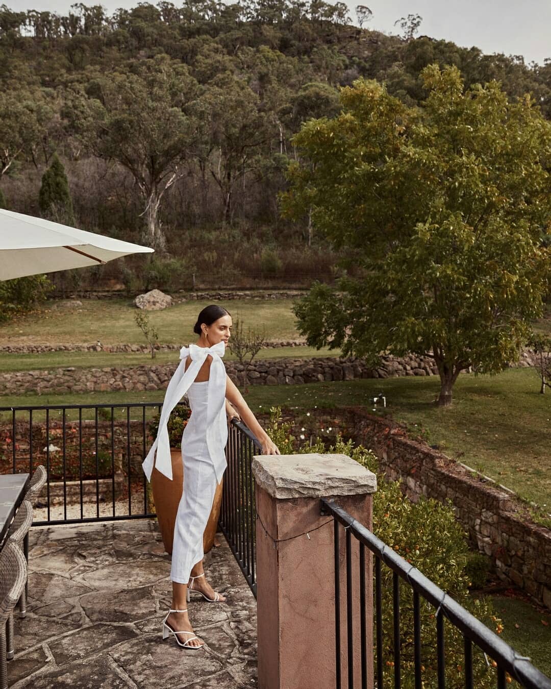 Woman wearing a white linen midi dress with long shoulder ties on a balcony