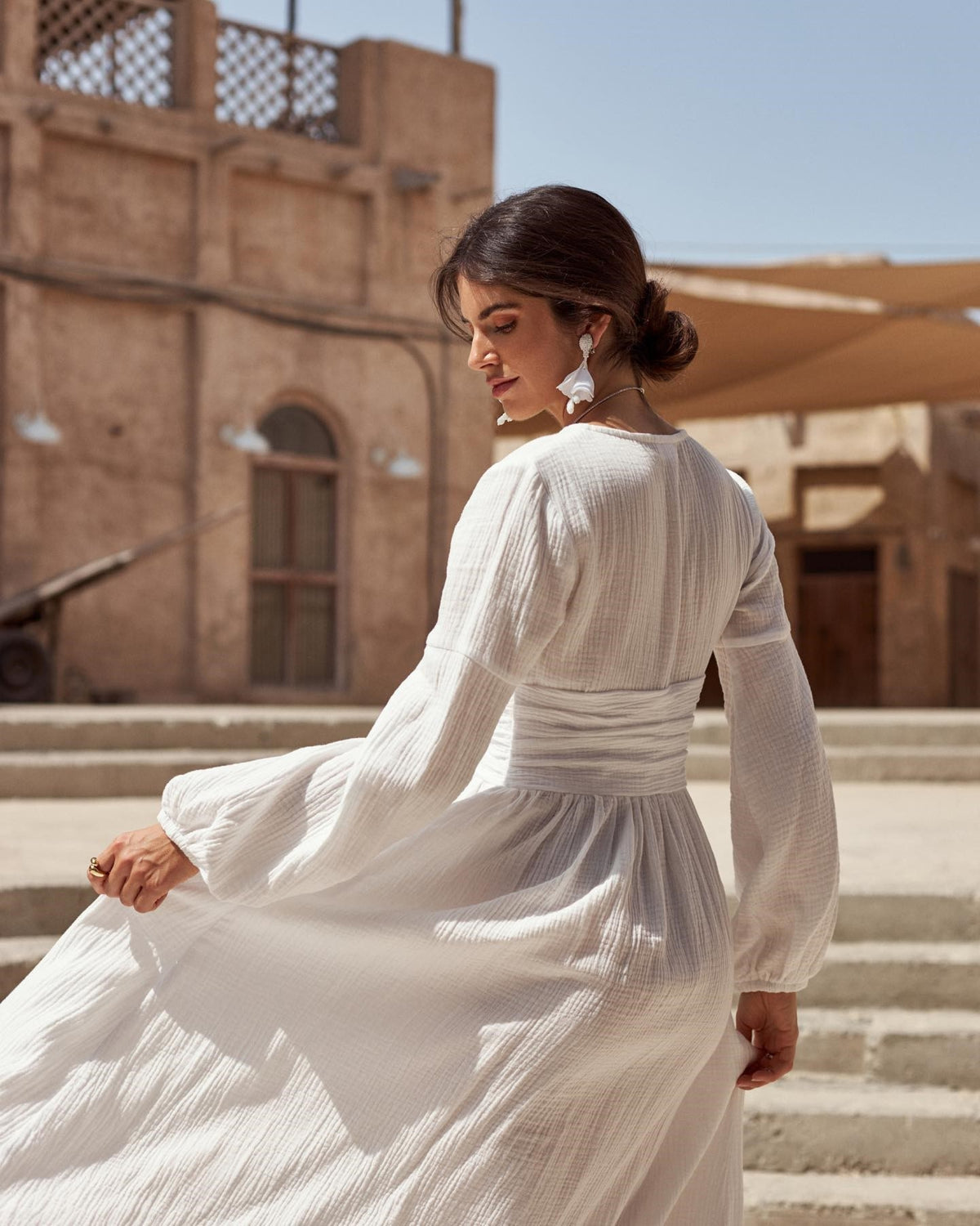 Back view of a white long sleeve muslin wedding dress with a defined draped waist and flowing floor-length skirt.