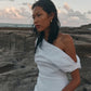 A woman sitting on a rocky beach at sunset, wearing a white one-shoulder linen dress.