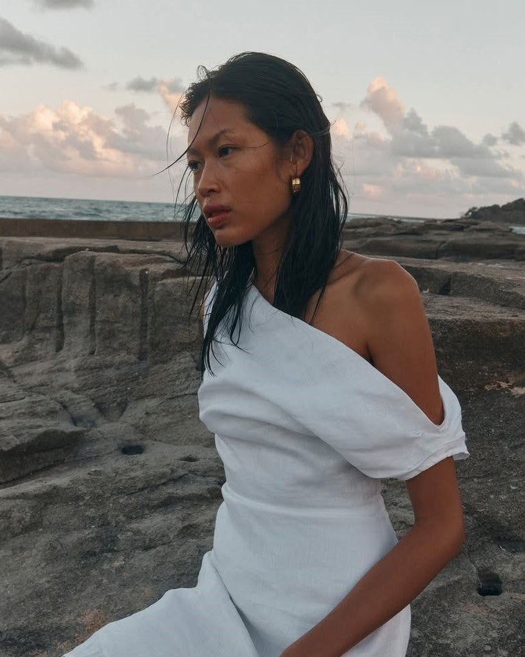A woman sitting on a rocky beach at sunset, wearing a white one-shoulder linen dress.