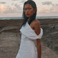 A woman stands on a seaside walkway at dusk wearing a white one-shoulder dress with a draped, oversized sleeve detail.