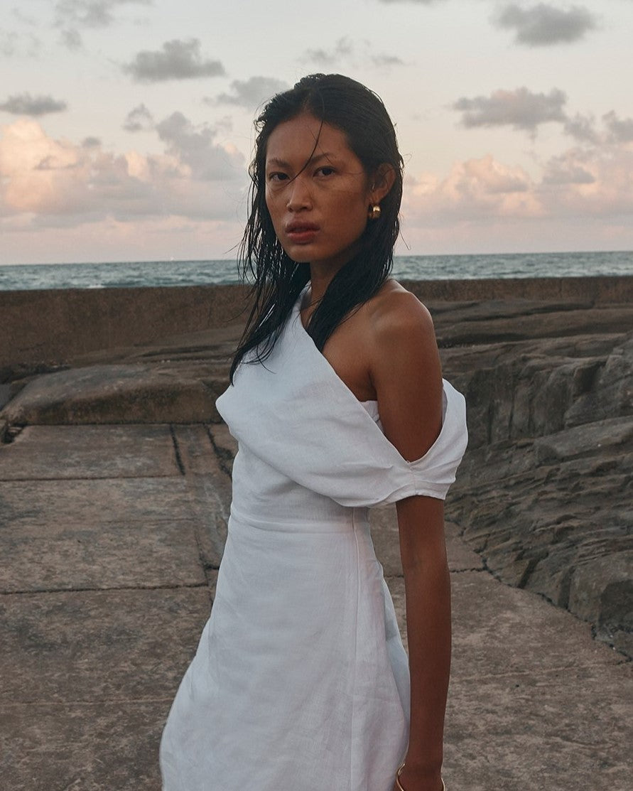 A woman stands on a seaside walkway at dusk wearing a white one-shoulder dress with a draped, oversized sleeve detail.