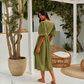 A woman wearing an olive green midi dress with puff sleeves and a tie waist, standing in a bright, modern outdoor patio.