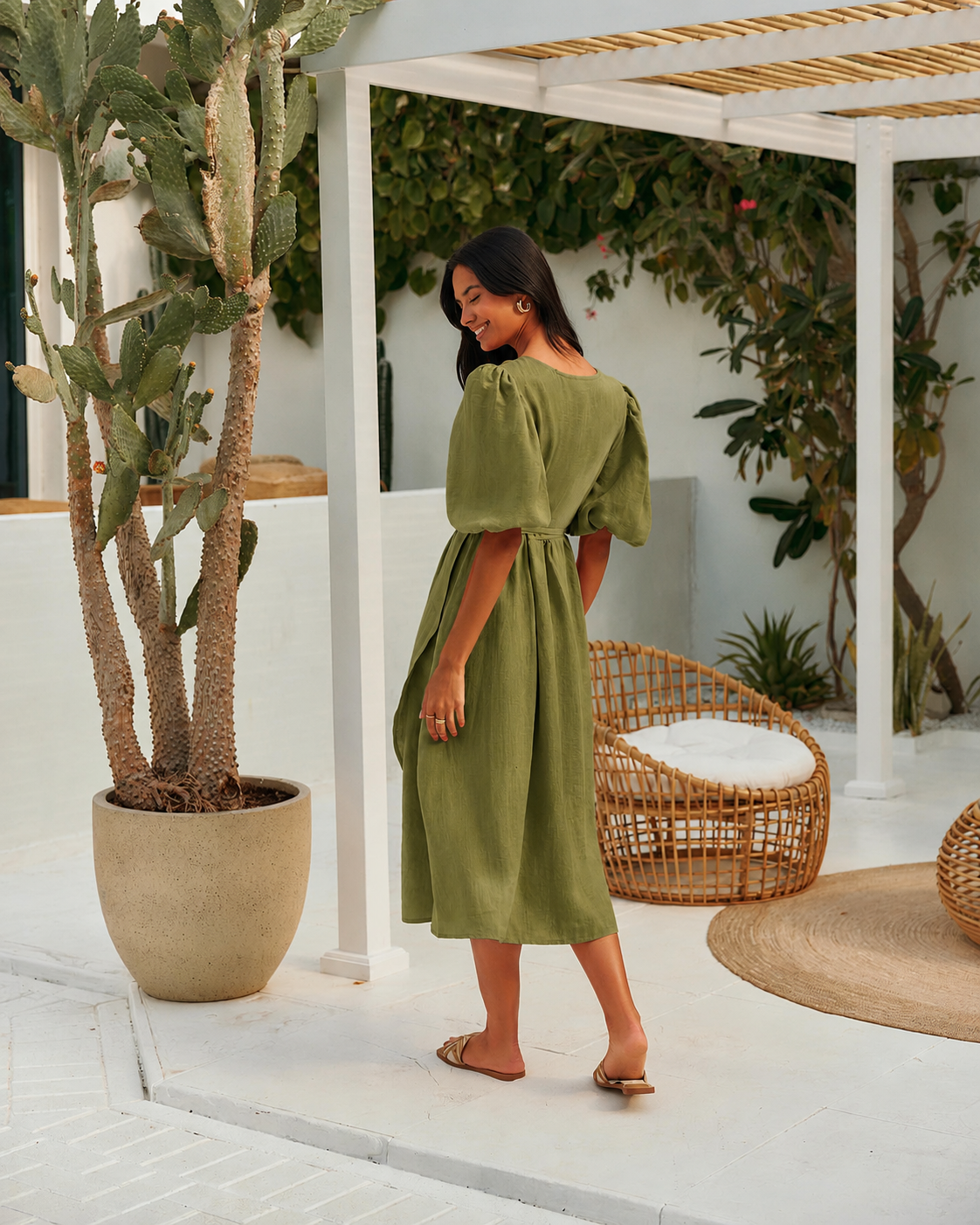 A woman wearing an olive green midi dress with puff sleeves and a tie waist, standing in a bright, modern outdoor patio.