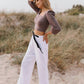 A person standing on a beach wearing wide-leg linen trousers with a contrasting belt, a light pink top, and white sandals.