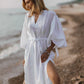 A woman standing on a beach wearing a white, loose-fitting maxi linen dress with a round neckline, balloon sleeves with tie detail at cuffs, and a wide drawstring waist. The dress has slits on the sides and is paired with a straw hat and a watch.