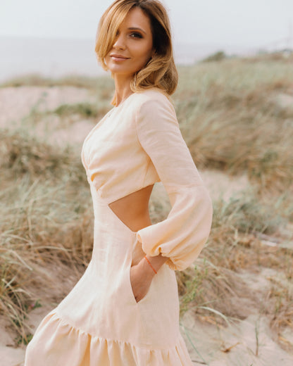 A woman wearing a peach A-line linen dress with 3/4 puff sleeves and an open back, standing in a natural outdoor setting.
