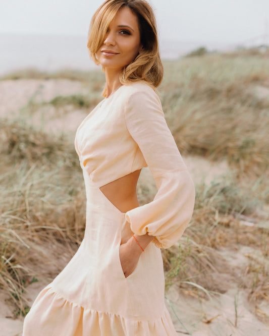 A woman wearing a peach A-line linen dress with 3/4 puff sleeves and an open back, standing in a natural outdoor setting.