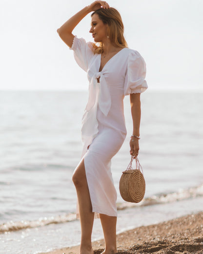A woman standing on a beach wearing a white midi linen dress with puff sleeves and an elastic cuff, holding a straw bag.