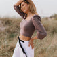 A woman standing on a beach wearing a lavender linen blouse with puff sleeves, square neckline, and wide straps, paired with white trousers.