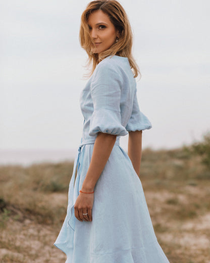 A woman wearing a long blue linen wrap dress with puff sleeves and a V-neckline, standing by the beach.