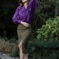 A woman standing in a forest setting, wearing a purple blouse with a round neckline and long sleeves, paired with a green plaid midi skirt with asymmetrical splits. The skirt has belt loops, and the outfit appears to be dry clean only.