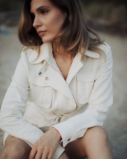 A person seated on a rocky surface wearing a white luxurious linen blend dress with a stand collar, shoulder epaulettes, long sleeves, and a front zipper closure, with no visible belt.