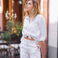 A woman wearing a white, oversized, long-sleeved linen shirt with patch pockets at the chest and long cuffed sleeves. She is also wearing white shorts, carrying a bag and wearing sandals.