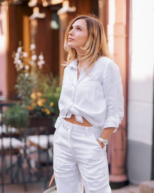 A woman wearing a white, oversized, long-sleeved linen shirt with patch pockets at the chest and long cuffed sleeves. She is also wearing white shorts, carrying a bag and wearing sandals.